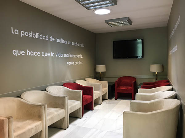Sala de espera con sillones de color crema y rojo, un televisor y una frase rotulada en la pared